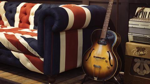 Eine alte akustische Gibson Gitarre steht auf einem Ständer neben einem Sofa mit dem Union Jack als Muster.