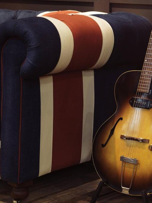 Eine alte akustische Gibson Gitarre steht auf einem Ständer neben einem Sofa mit dem Union Jack als Muster.