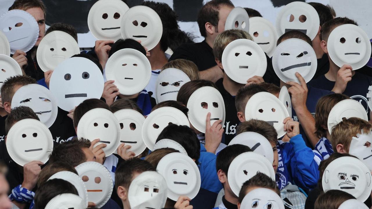 Karlsruher Fans protestieren gegen Gesichtserkennung im Stadion, indem sie sich Pappteller mit gemalten Gesichtern vor das Gesicht halten