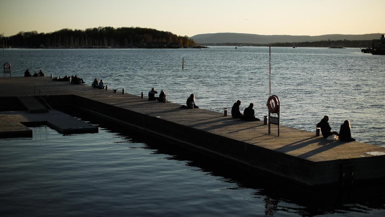 Auf einem Bootssteg, der ins Wasser ragt, sitzen mehrere Pärchen mit deutlichem Abstand voneinander. Auf einem Bootssteg, der ins Wasser ragt, sitzen mehrere Pärchen mit deutlichem Abstand voneinander.