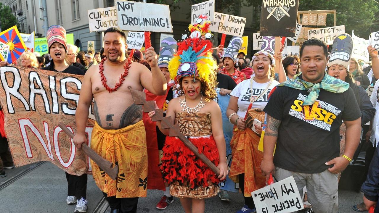 Man sieht auf einer Straße eine Gruppe von Demonstranten von den pazifischen Inseln, die Plakate und Schilder für einen besseren Klimaschutz halten. Einige tragen traditioneller Kleidung. Man sieht auf einer Straße eine Gruppe von Demonstranten von den pazifischen Inseln, die Plakate und Schilder für einen besseren Klimaschutz halten. Einige tragen traditioneller Kleidung.