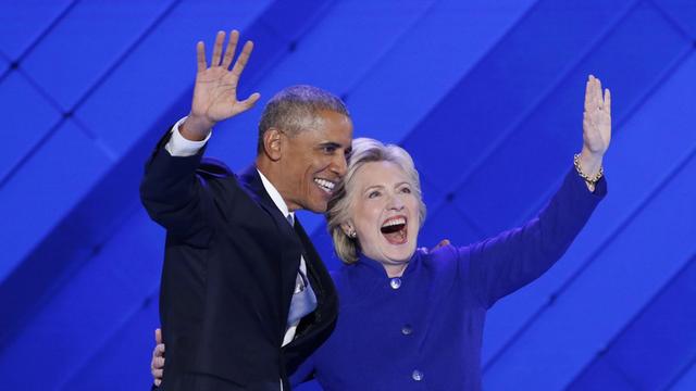 Clinton und Obama stehen gemeinsam auf der Bühne. Sie lachen und winken. 