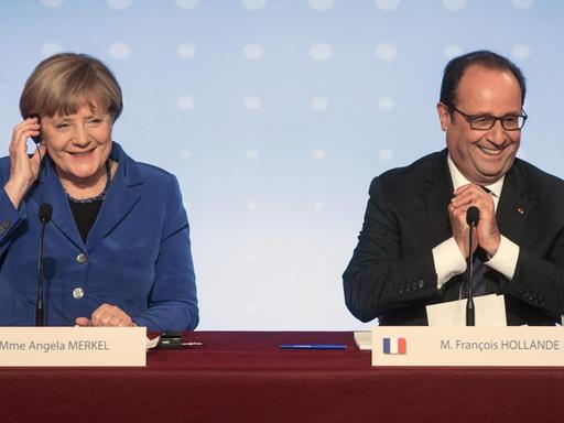 Bundeskanzlerin Angela Merkel und Frankreichs Präsident Francois Hollande geben nach dem Ukraine-Gipfel eine Pressekonferenz. Sie sitzen hinter einem Pult und grinsen.