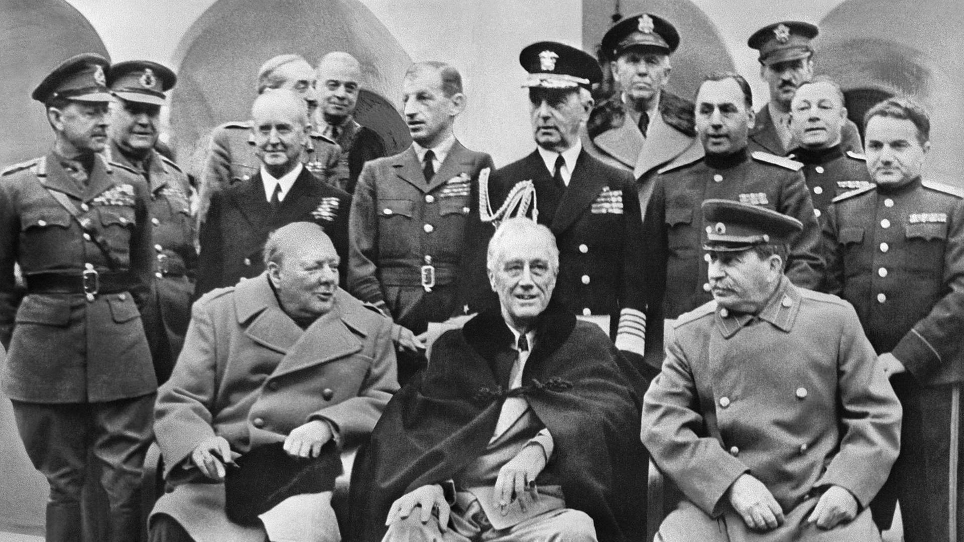 Eine historische Aufnahme zeigt den den britischen Prämierminister Winston Churchill zusammen mit US-Präident Franklin D. Roosevelt und Joseph Stalin bei der Konferenz in Jalta am 11.02.1945 sitzend. Im Hintergrund stehen zwahlreiche Männer in militärischen Uniformen.