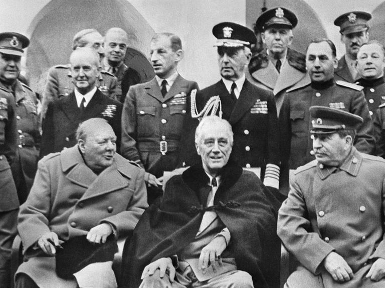 Eine historische Aufnahme zeigt den den britischen Prämierminister Winston Churchill zusammen mit US-Präident Franklin D. Roosevelt und Joseph Stalin bei der Konferenz in Jalta am 11.02.1945 sitzend. Im Hintergrund stehen zwahlreiche Männer in militärischen Uniformen.