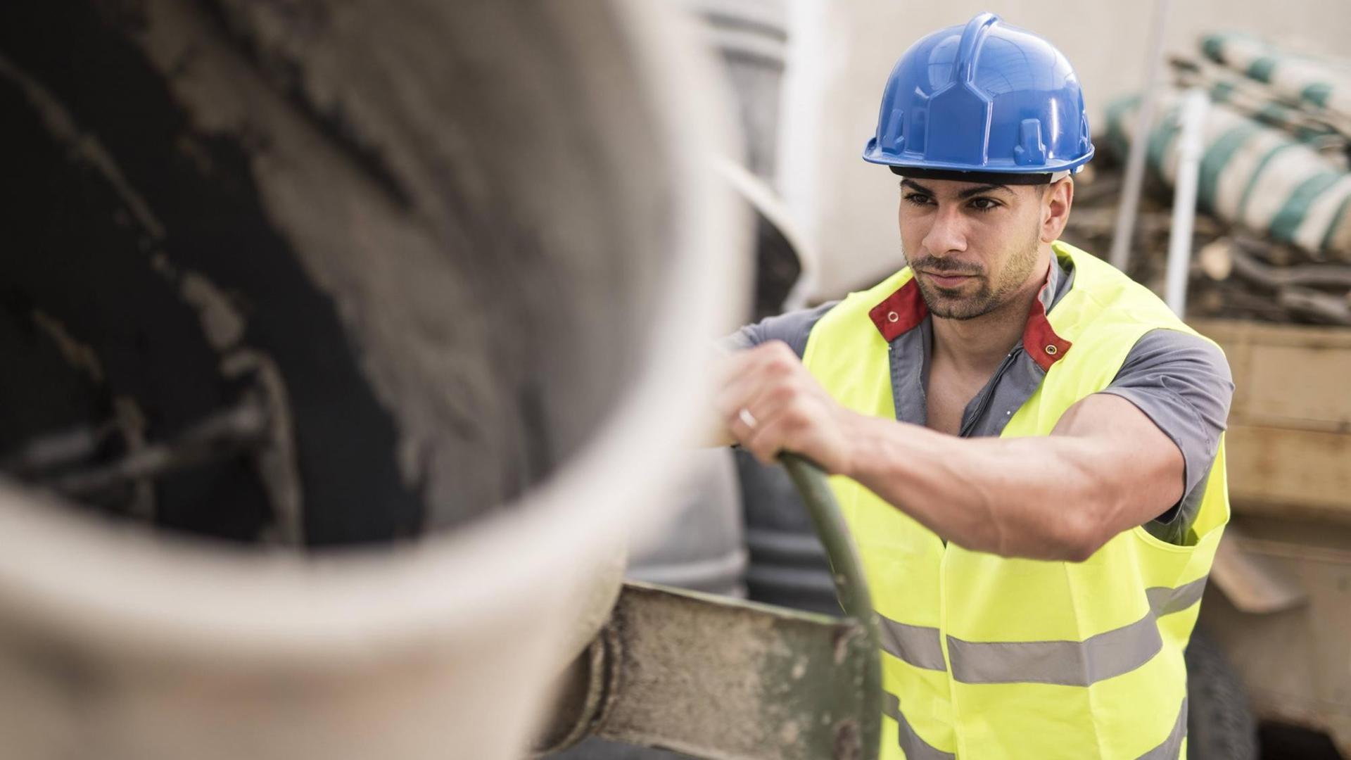 Ein junger Bauarbeiter steht an einer Betonmischmaschine auf einer Baustelle. Er trägt eine gelbe Warnweste und einen bleuen Helm. Ein junger Bauarbeiter steht an einer Betonmischmaschine auf einer Baustelle. Er trägt eine gelbe Warnweste und einen bleuen Helm.