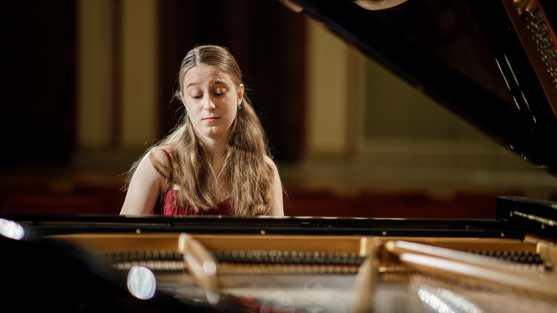 Die junge Pianistin ist vom Flügelinneren aus fotografiert. Sie hat lange, hellbraune Haare und trägt ein ärmelloses rotes Kleid, sie blickt beim Spielen auf die Tasten, der Deckel des schwarzen Flügels ist aufgeklappt. Die junge Pianistin ist vom Flügelinneren aus fotografiert. Sie hat lange, hellbraune Haare und trägt ein ärmelloses rotes Kleid, sie blickt beim Spielen auf die Tasten, der Deckel des schwarzen Flügels ist aufgeklappt.