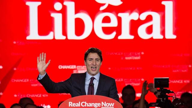 Justin Trudeau steht mit erhobenen Armen vor einer Wahl-Werbubg seiner Partei und hält eine Rede.