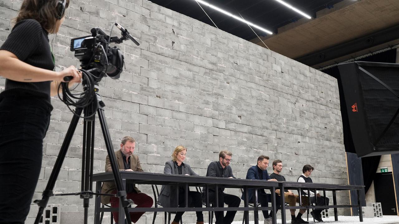 Sechs Schauspieler sitzen an einem langen Tisch im Hellen, schauen auf ihre Handys und werden gefilmt.