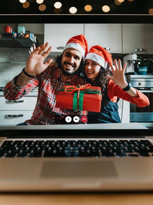 Ein junges Paar mit Weihnachtsmützen auf dem Kopf und Geschenk in der Hand winkt in die Kamera eines Laptop-Bildschirms.