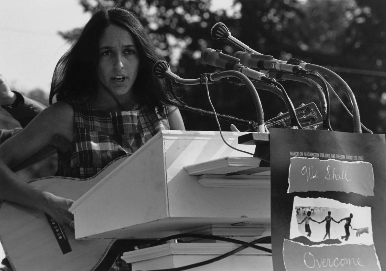 Eine junge Sängerin steht hinter einem großen Pult mit vielen Mikrophonen und spielt Gitarre, während ein Schild am Pult den Schriftzug "We Shall Overcome" zeigt. Eine junge Sängerin steht hinter einem großen Pult mit vielen Mikrophonen und spielt Gitarre, während ein Schild am Pult den Schriftzug "We Shall Overcome" zeigt.
