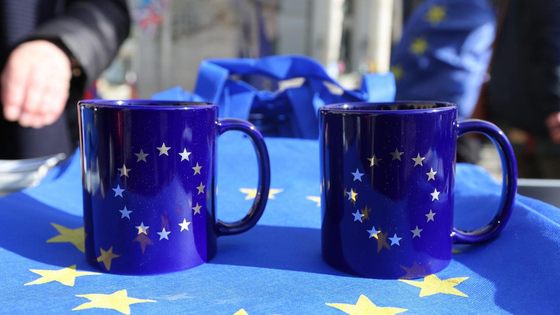 Zwei Tassen mit Europa-Flaggen-Aufdruck stehen auf einem Tisch, der von einer Europaflagge bedeckt ist