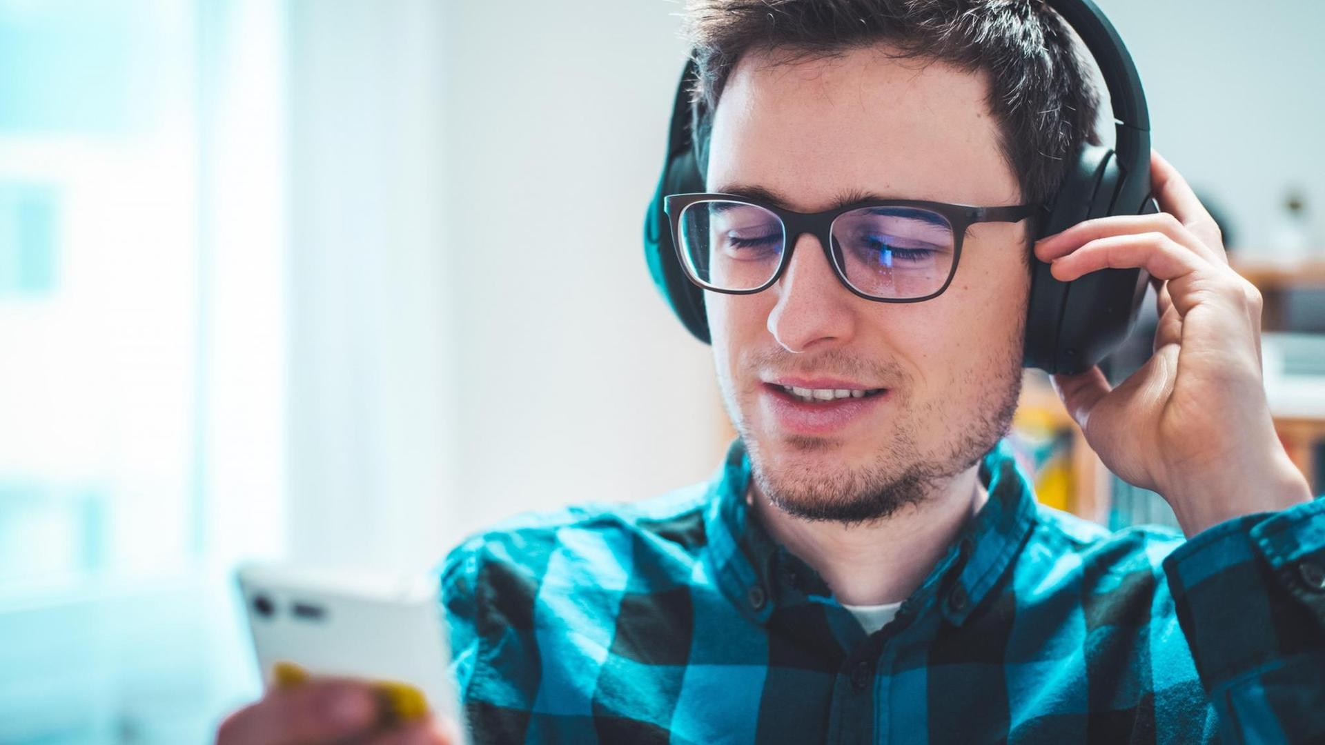 Ein junger weißer Mann in kariertem Hemd und mit Brille hört mit Kopfhörern etwas und schaut dabei auf sein Smartphone. Ein junger weißer Mann in kariertem Hemd und mit Brille hört mit Kopfhörern etwas und schaut dabei auf sein Smartphone.