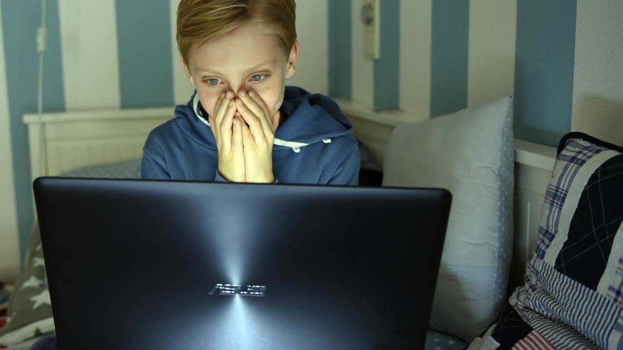 Ein Junge sitzt mit einem Laptop auf seinem Bett, hat die Hände vor das Gesicht geschlagen und schaut sich einen Horrorfilm an. Ein Junge sitzt mit einem Laptop auf seinem Bett, hat die Hände vor das Gesicht geschlagen und schaut sich einen Horrorfilm an.