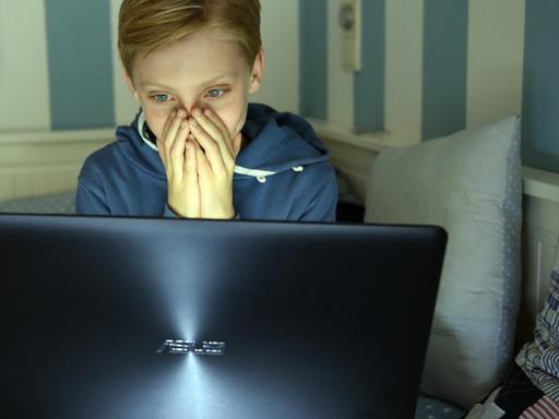 Ein Junge sitzt mit einem Laptop auf seinem Bett und hat die Hände vor das Gesicht geschlagen.