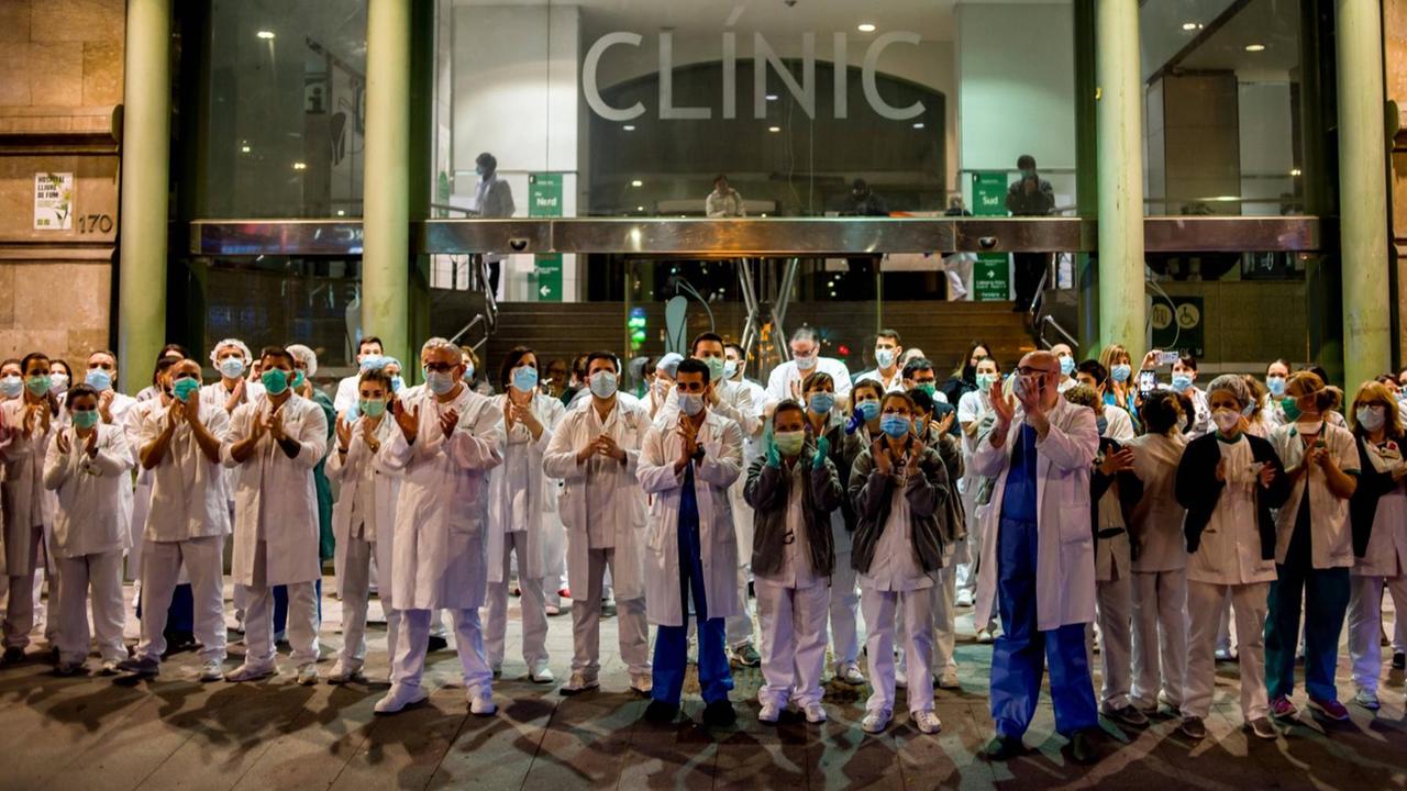 Klinikmitarbeiter in Kitteln und mit Mundschutz stehen vor dem Krankenhaus auf der Straße und applaudieren. Klinikmitarbeiter in Kitteln und mit Mundschutz stehen vor dem Krankenhaus auf der Straße und applaudieren.