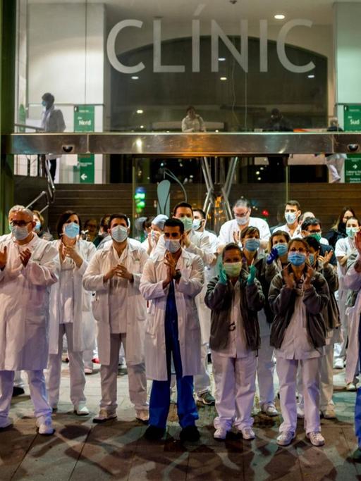 Klinikmitarbeiter in Kitteln und mit Mundschutz stehen vor dem Krankenhaus auf der Straße und applaudieren.