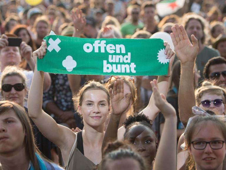 Demonstranten bei der Abschlusskundgebung der #unteilbar-Demo in Dresden. Eine lächelnde junge Frau hält inmitten einer heiteren Menschenmenge ein Plakat mit der Aufschrift "offen und bunt" in die Höhe.