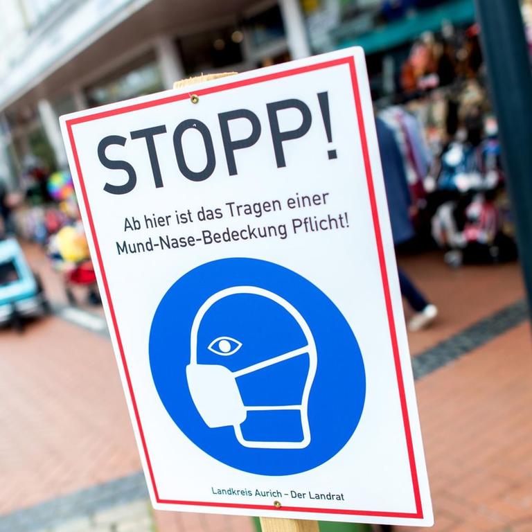 Ein Schild mit der Aufschrift "Stopp! Ab hier ist das Tragen einer Mund-Nasen-Bedeckung Pflicht!" steht in einer Fußgängerzone.