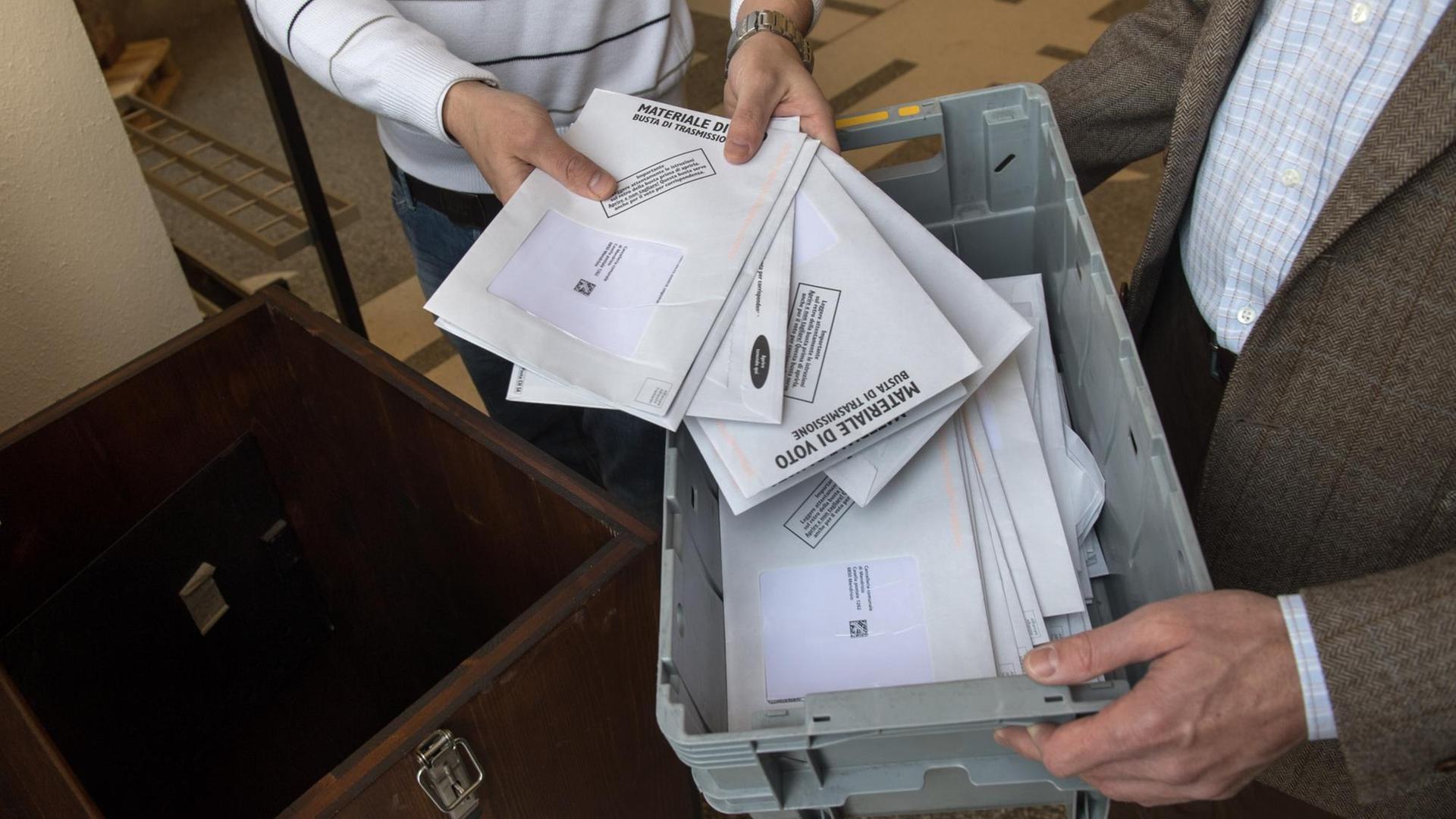 Zwei Hände halten einen Plastikkorb, zwei andere holen Wahlbriefe heraus.