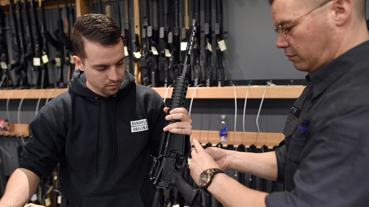 Die beiden Verkäufer sind schwarz gekleidet und hängen ein Schild an ein Gewehr. Im Hintergrund sieht man in einer Wandhalterung zahlreiche weitere Gewehre. Die beiden Verkäufer sind schwarz gekleidet und hängen ein Schild an ein Gewehr. Im Hintergrund sieht man in einer Wandhalterung zahlreiche weitere Gewehre.