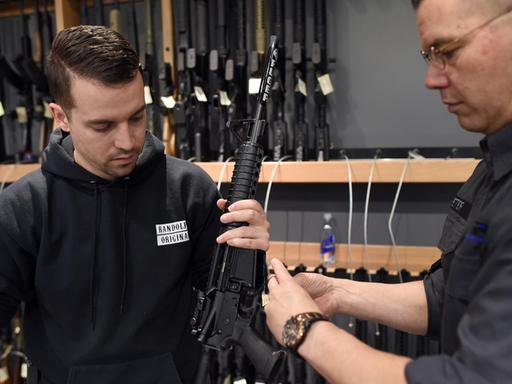 Die beiden Verkäufer sind schwarz gekleidet und hängen ein Schild an ein Gewehr. Im Hintergrund sieht man in einer Wandhalterung zahlreiche weitere Gewehre.