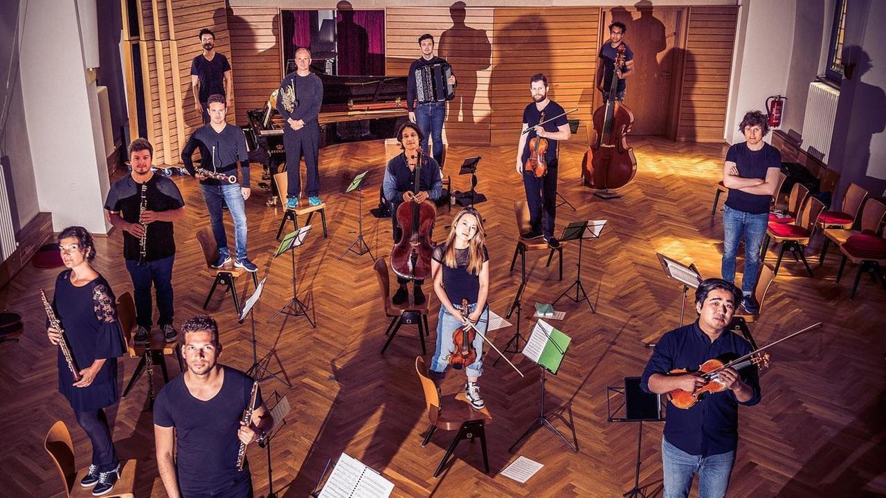 Etliche Musiker stehen in Sicherheitsabstand voneinander in einem Probenraum mit gepflegtem Parkett. Etliche Musiker stehen in Sicherheitsabstand voneinander in einem Probenraum mit gepflegtem Parkett.