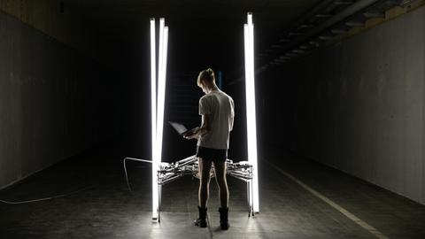 Das Foto zeigt ein Kunstwerk auf der Ars Electronica: Es besteht unter anderem aus senkrecht aufgestellten, hell erleuchteten Neonröhren in einem dunklen Raum. Die Röhren sind durch eine technsiche Konstruktion miteinander verbunden.