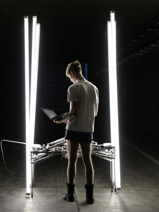 Das Foto zeigt ein Kunstwerk auf der Ars Electronica: Es besteht unter anderem aus senkrecht aufgestellten, hell erleuchteten Neonröhren in einem dunklen Raum. Die Röhren sind durch eine technsiche Konstruktion miteinander verbunden.