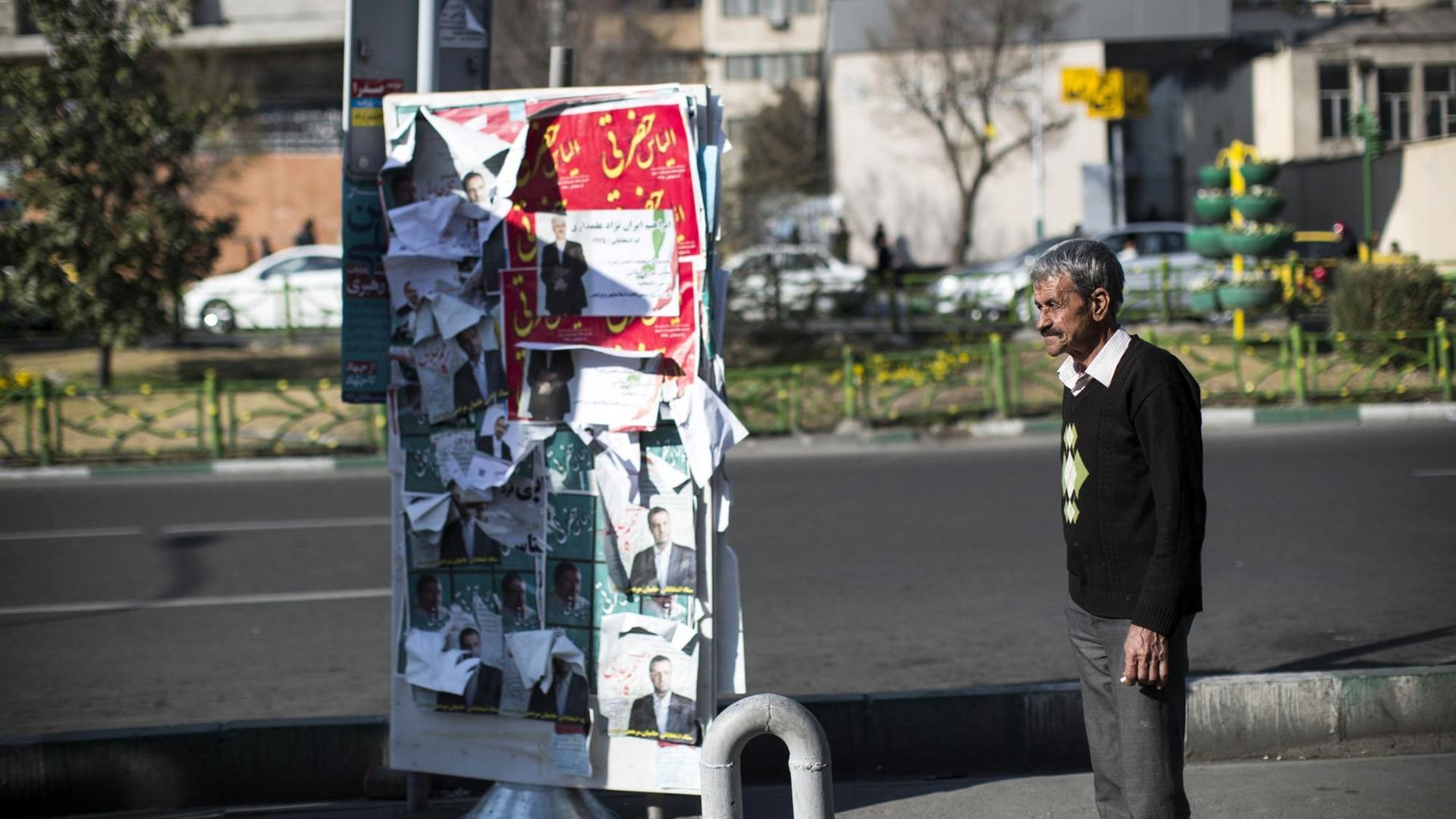 Ein Mann steht in Teheran vor einer Säule mit Wahlplakaten.