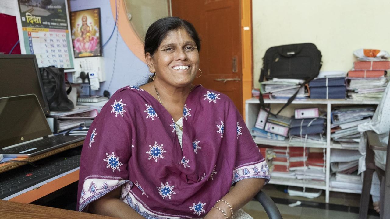 Eine fröhlich lächelnde Frau - das schwarze Haar zum langen Zopf gebunden -  sitzt in einem lila Sari in ihrem vollgepackten Büro am Schreibtisch.