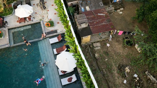 Reich und Arm in Vietnam dicht beieinander: Eine moderne Hotelanlage mit Schwimmbecken und grünen Pflanzen in Hoi An ist nur durch eine weiße Mauer getrennt von einem verrosteten Haus mit rostigem Wellblechdach.
