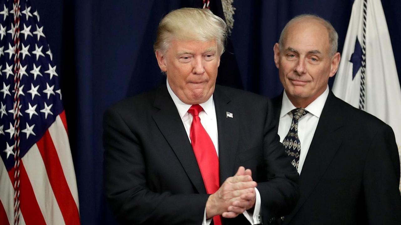 Trump steht zwischen einer US-Flagge und einer anderen Flagge und reibt sich die Hände; Kelly steht lächelnd dahinter. Trump steht zwischen einer US-Flagge und einer anderen Flagge und reibt sich die Hände; Kelly steht lächelnd dahinter.