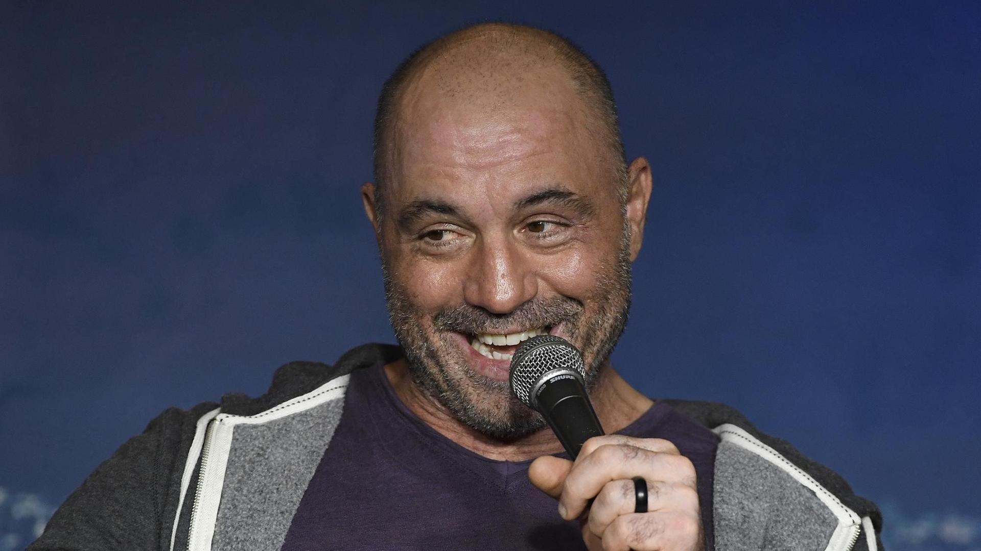 Das Bild zeigt den US-amerikanischen Comedia Joe Roegan, der mit seinem Podcast "The Joe Rogan Experience" berühmt geworden ist. Im Bild sitzt er mit kurzgeschorenen Haaren und breitem Lächeln mit einem Mikrofon in der Hand auf einer Bühne. Das Bild zeigt den US-amerikanischen Comedia Joe Roegan, der mit seinem Podcast "The Joe Rogan Experience" berühmt geworden ist. Im Bild sitzt er mit kurzgeschorenen Haaren und breitem Lächeln mit einem Mikrofon in der Hand auf einer Bühne.