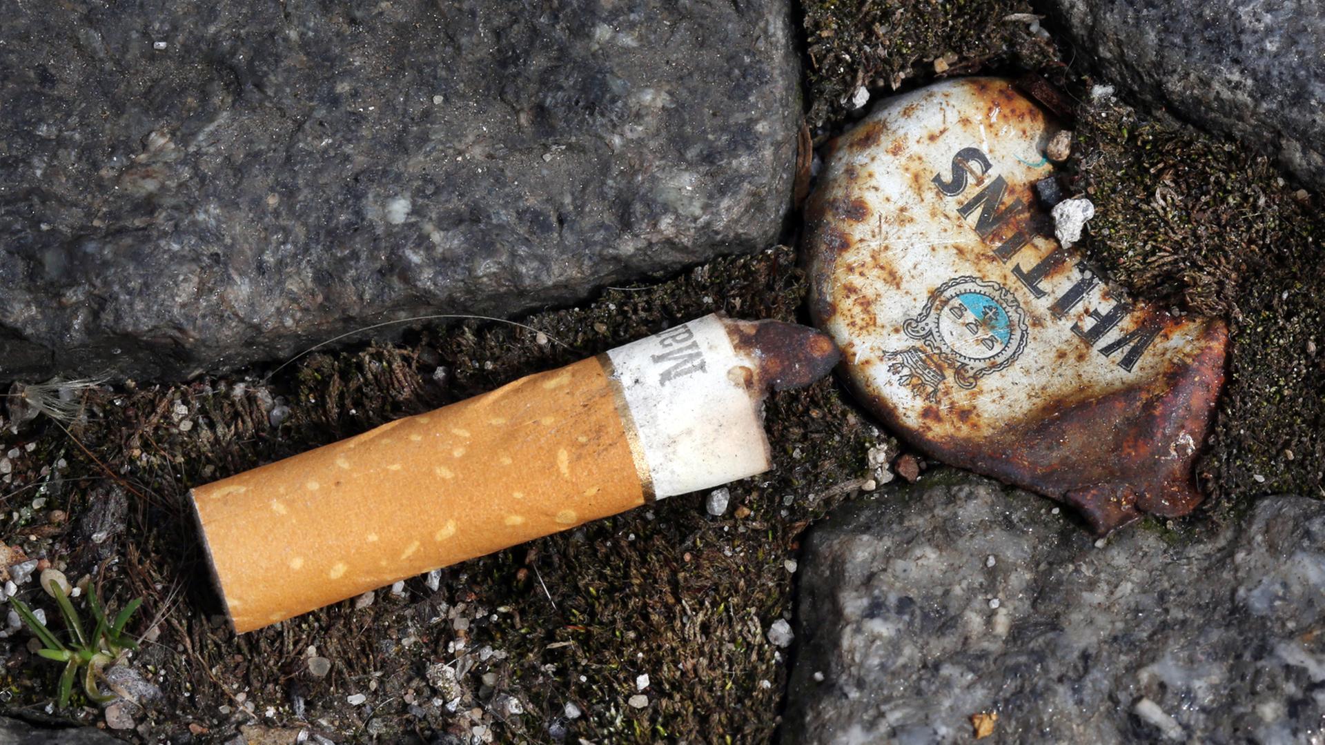 Ein Zigarettenstummel liegt neben einem rostigen Kronkorken zwischen Pflastersteinen auf dem Boden.