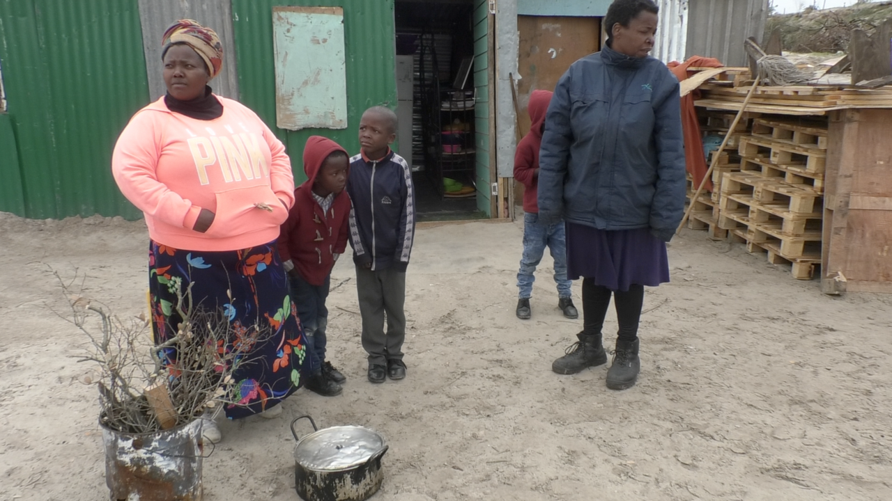 Zoleka Velengceni mit drei Kindern und einer Frau vor einer Wellblechhütte. Ein Topf mit Deckel und ein Eimer mit Zweigen stehen auf dem Boden. Zoleka Velengceni mit drei Kindern und einer Frau vor einer Wellblechhütte. Ein Topf mit Deckel und ein Eimer mit Zweigen stehen auf dem Boden.