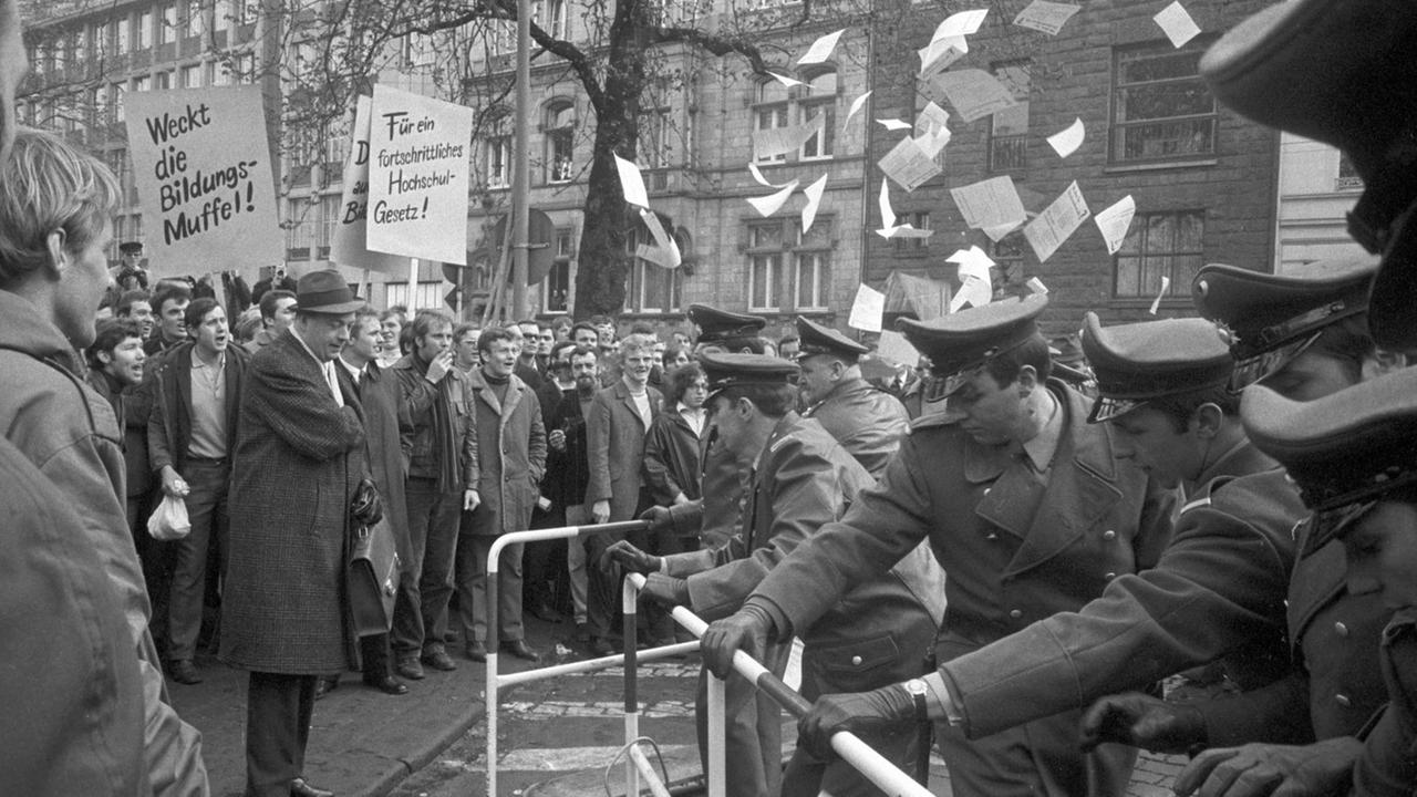 Studierende demonstrieren im April 1969 mit Plakaten und Flyern. Gegenüber stehen ihnen uniformierte Beamte hinter einem Zaun. Studierende demonstrieren im April 1969 mit Plakaten und Flyern. Gegenüber stehen ihnen uniformierte Beamte hinter einem Zaun.