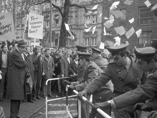 Studierende demonstrieren im April 1969 mit Plakaten und Flyern. Gegenüber stehen ihnen uniformierte Beamte hinter einem Zaun.