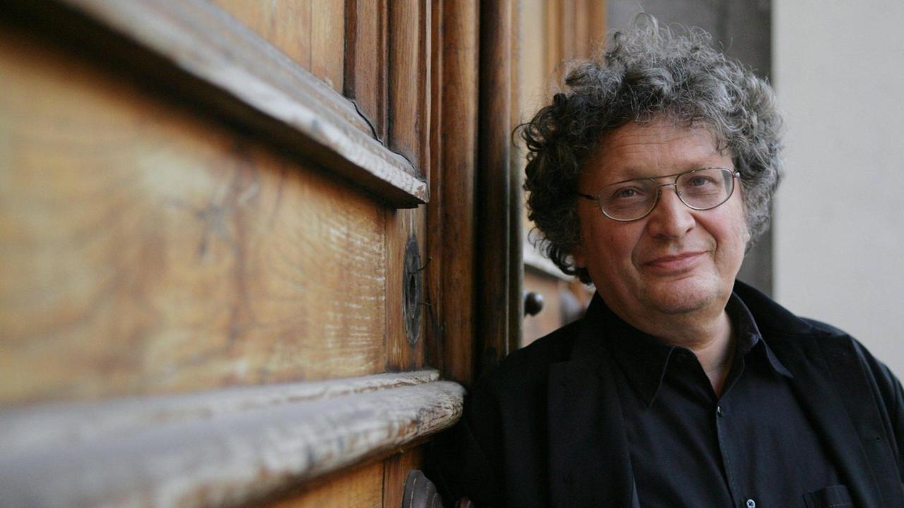 Der Dirigent mit grau-schwarzen Locken und Brille lehnt an einer schweren, historischen Holztür und lächelt in die Kamera. Der Dirigent mit grau-schwarzen Locken und Brille lehnt an einer schweren, historischen Holztür und lächelt in die Kamera.