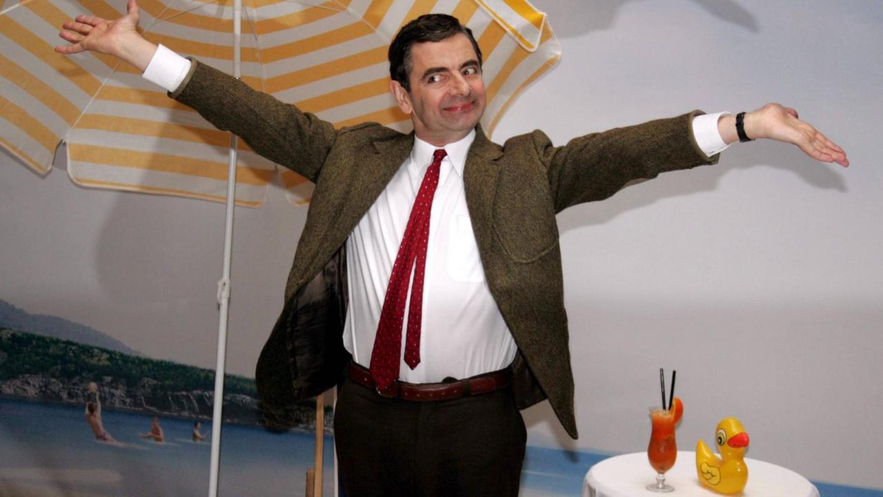 Komiker Rowan Sebastian Atkinson (GBR) anlässlich eines Pressetermins zu seinem Film - Mr. Bean macht Ferien - in Berlin: Er steht vor einer Karibiktapete und einem Sonnenschirm und hält die Hände in die Höhe.