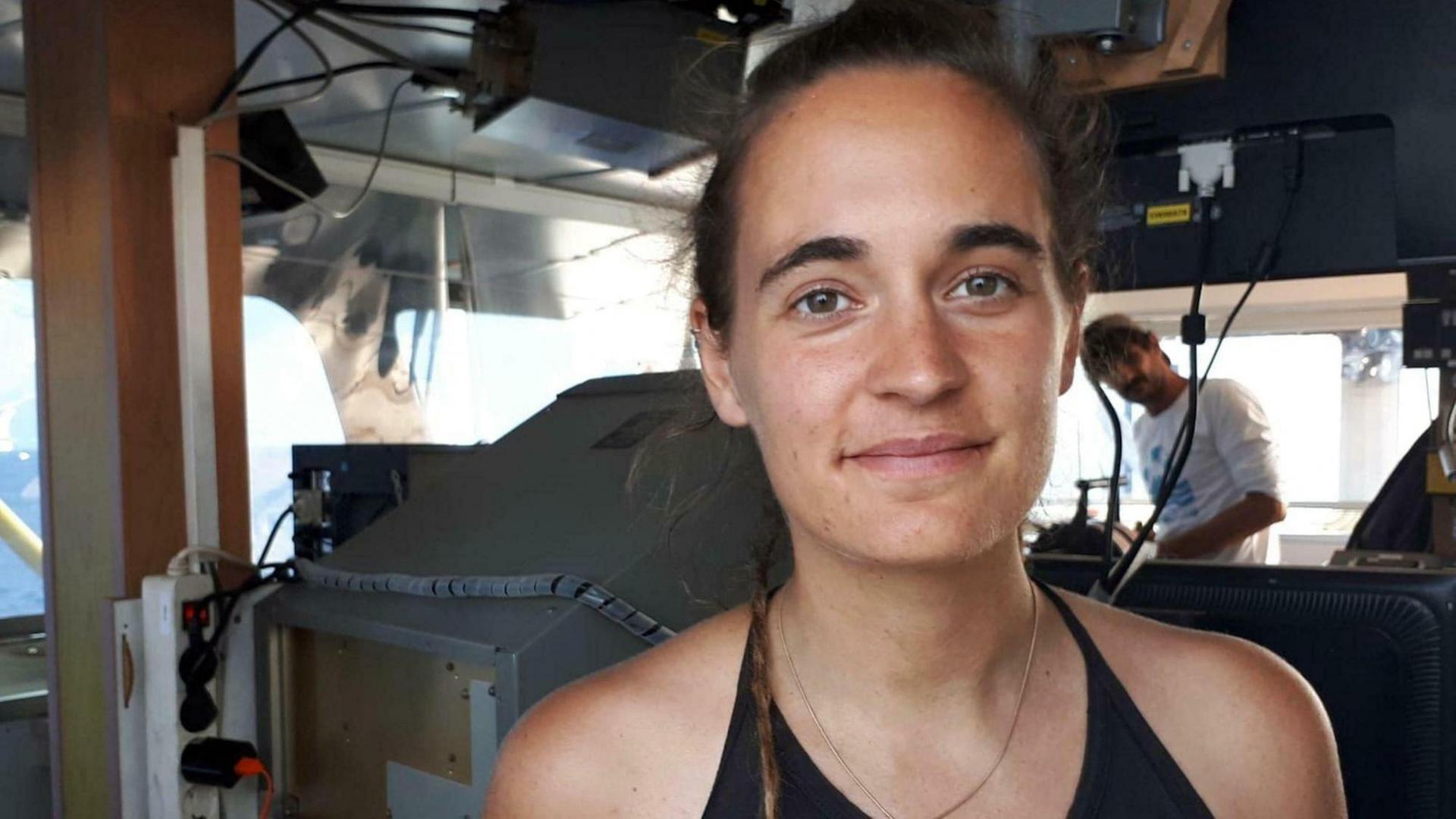 Kapitänin Carola Rackete steht auf der Brücke ihres Schiffes und lächelt in die Kamera. Dahinter unscharf ein Mann. Kapitänin Carola Rackete steht auf der Brücke ihres Schiffes und lächelt in die Kamera. Dahinter unscharf ein Mann.