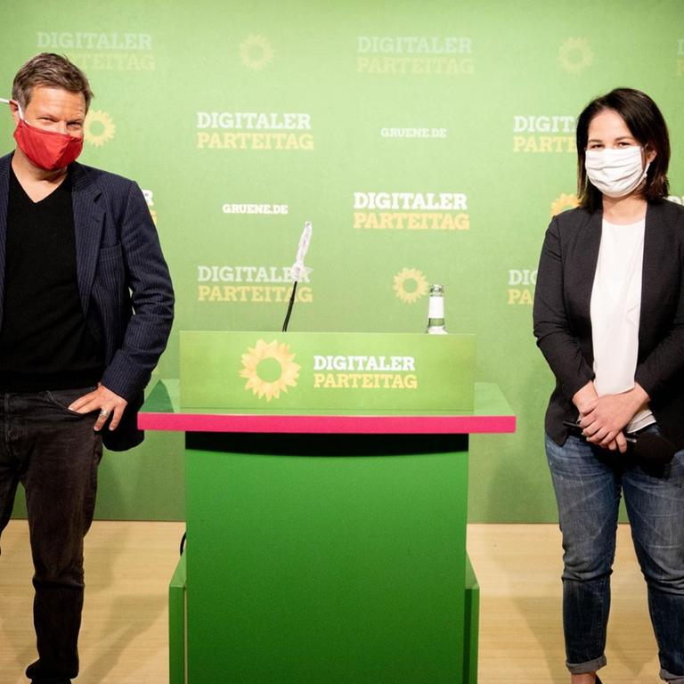 Die Grünen-Bundesvorsitzenden Robert Habeck (l) und Annalena Baerbock (r) auf dem Parteitag, der wegen der Corona-Pandemie nur im Internet übertragen wird. Sie stehen vor einer Wand mit dem Logo der Grünen, halten Abstand voneinander und tragen Mund-Nasen-Masken.