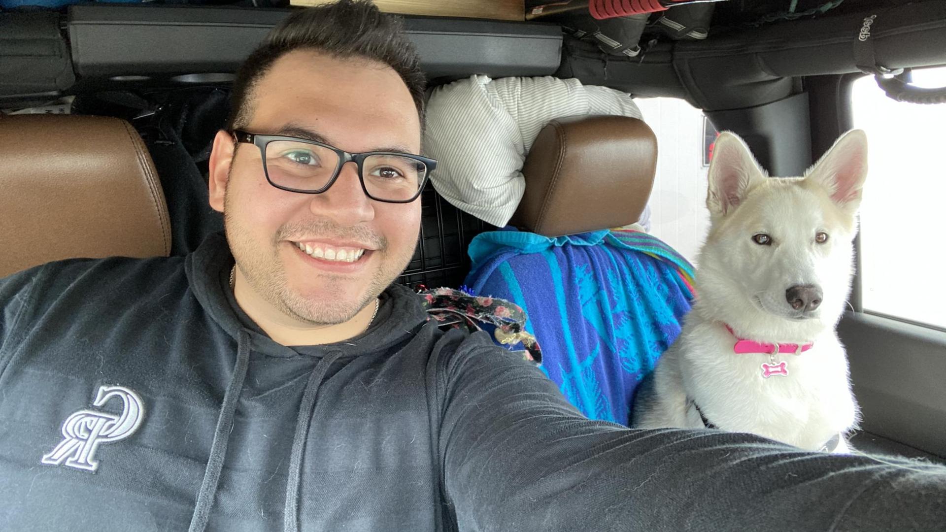 Ein lächelnder junger Mann, Oscar Garza, mit Brille sitzt im Auto. Neben ihm eine weiße Hündin auf dem Beifahrersitz. Ein lächelnder junger Mann, Oscar Garza, mit Brille sitzt im Auto. Neben ihm eine weiße Hündin auf dem Beifahrersitz.