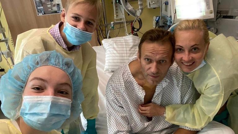 Die Nawalny-Kinder und seine Frau tragen gelbe Schutzkittel, die Kinder auch Mundschutz. Alle grinsen in die Kamera, nur Nawalny im Krankenhauskittel blickt müde. Seine Frau umarmt ihn.