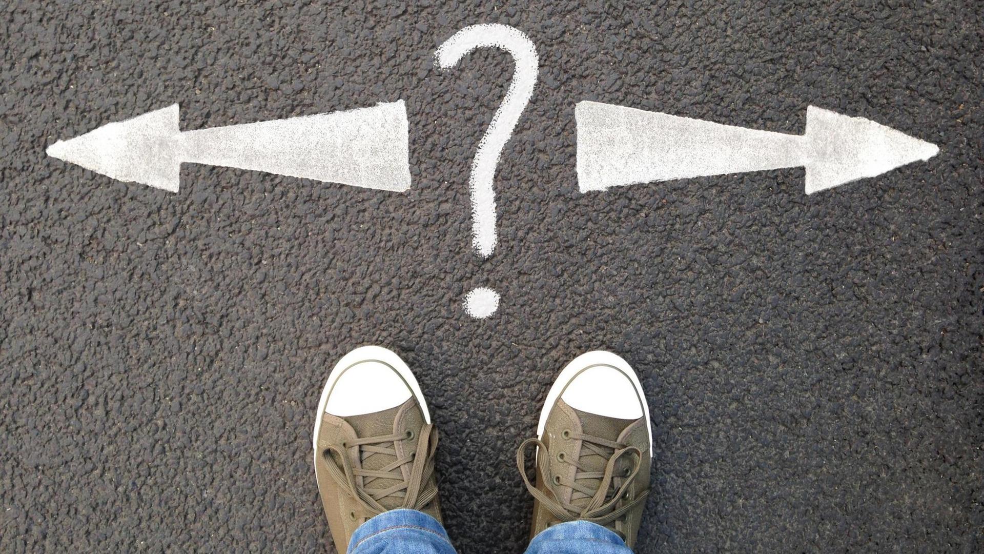 Füße in Turnschuhen stehen vor einer Markierung auf dem Asphalt: ein Fragezeichen und zwei Pfeile in entgegengesetzte Richtungen. (Symbolfoto) Füße in Turnschuhen stehen vor einer Markierung auf dem Asphalt: ein Fragezeichen und zwei Pfeile in entgegengesetzte Richtungen. (Symbolfoto)