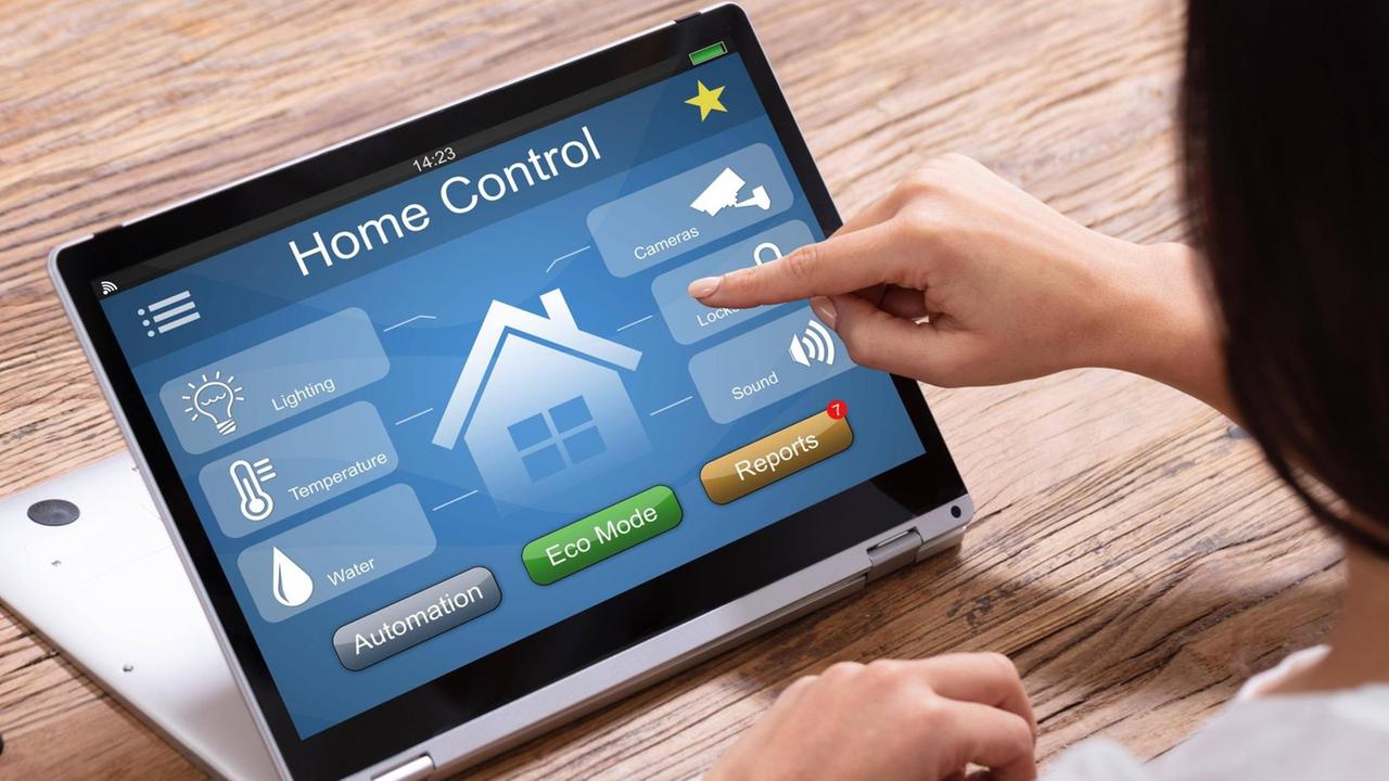 Eine Frau bedient an einem Tablet mit Touchscreen ein Smart Home Control System. Auf dem Bildschirm sind Piktogramme zu sehen, die für die einzelnen Bereiche des Hauses stehen, die man kontrollieren kann: Licht, Temperatur, Wasser, Überwachungskameras und Schließmechanismen.