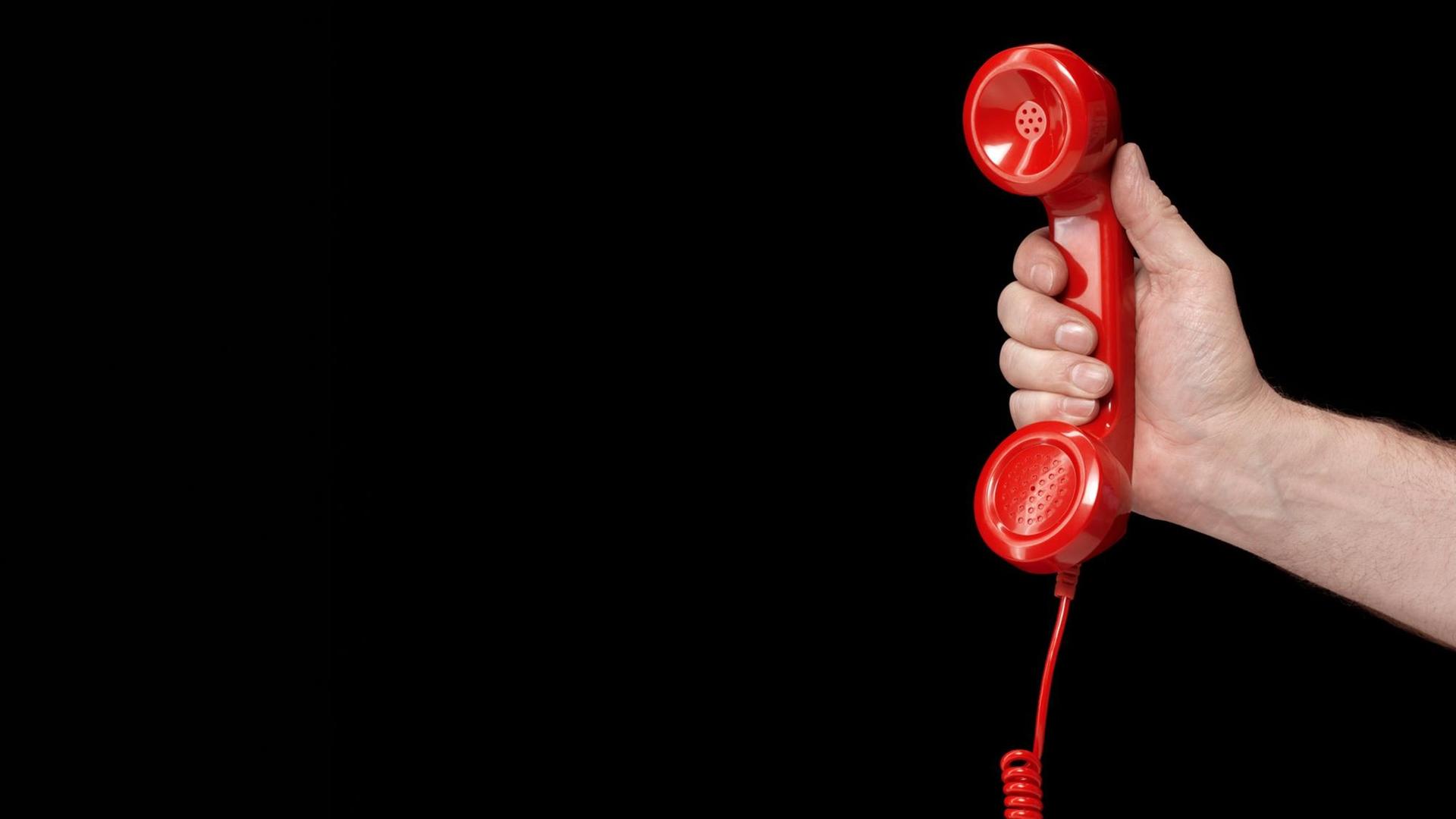 Eine Hand hält einen roten Telefonhörer vor schwarzem Hintergrund. Eine Hand hält einen roten Telefonhörer vor schwarzem Hintergrund.