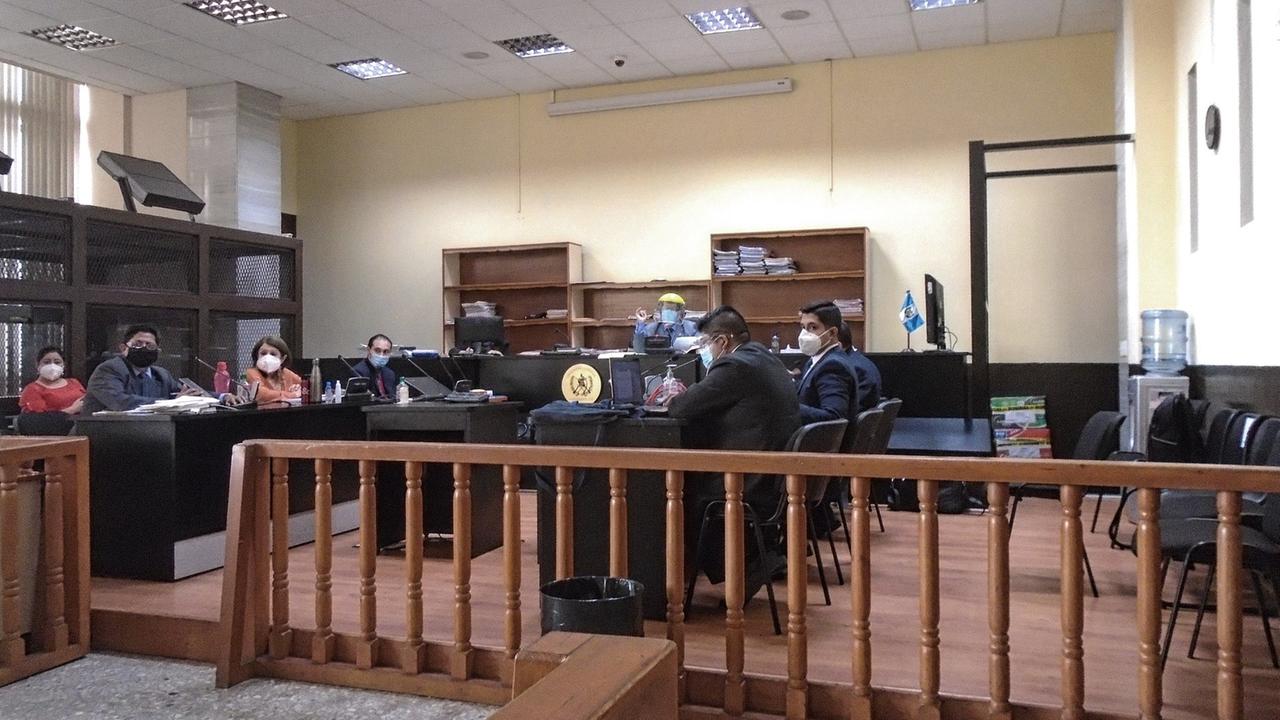 Blick in einen schlichten Amtsraum, hinter einem Holzgeländer sitzen 7 Personen einander an zwei langen Tischen gegenüber, im Hintergrund sitzt ein Mann erhöht am Richterpult, alle Tragen wegen der weltweiten Covid-19 Pandemie Mund-Nase-Masken. Blick in einen schlichten Amtsraum, hinter einem Holzgeländer sitzen 7 Personen einander an zwei langen Tischen gegenüber, im Hintergrund sitzt ein Mann erhöht am Richterpult, alle Tragen wegen der weltweiten Covid-19 Pandemie Mund-Nase-Masken.