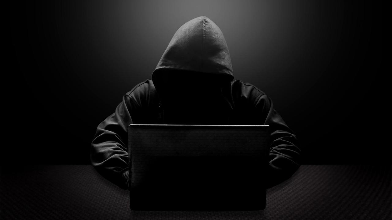 Ein Mann mit schwarzer Kapuze und verschattetem Gesicht sitzt vor einem Laptop.