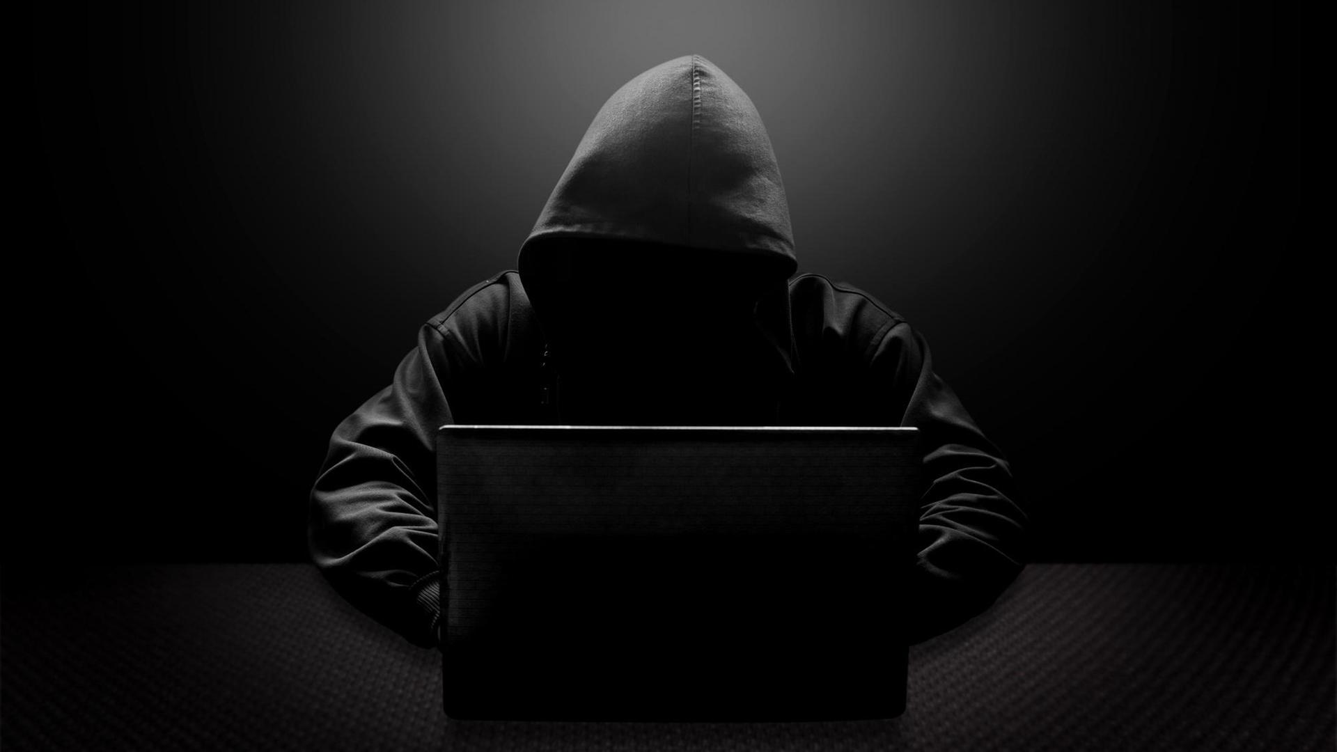 Ein Mann mit schwarzer Kapuze und verschattetem Gesicht sitzt vor einem Laptop. Ein Mann mit schwarzer Kapuze und verschattetem Gesicht sitzt vor einem Laptop.